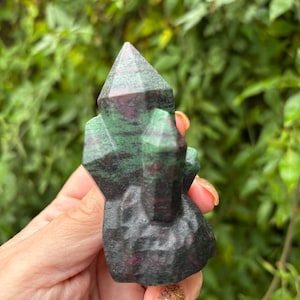 May include: A carved gemstone sculpture featuring a cluster of green and red crystals. The crystals have a faceted design and are set on a dark base. The sculpture is held against a backdrop of green foliage.