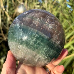 May include: A green, white and purple fluorite sphere with a smooth, polished surface. The sphere is approximately 5 cm in diameter and has a natural, organic look.