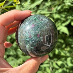 May include: A polished gemstone sphere, mainly green with red and black inclusions. The sphere is held in a hand, with a blurred green background. The surface of the sphere reflects the surrounding environment.