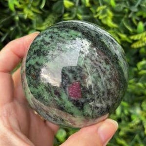 May include: Green and black gemstone sphere with red inclusions. The sphere is polished and has a smooth surface.