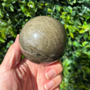 May include: A polished, spherical, golden sheen obsidian gemstone. The orb is held in a hand, with a backdrop of green foliage. The stone has a smooth, reflective surface, showcasing natural patterns and colour variations.