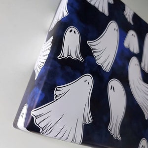 May include: A roll of wrapping paper with a dark blue background and a pattern of white cartoon ghosts. The ghosts have simple, rounded shapes and are scattered across the paper. The paper is partially unrolled, revealing the pattern.