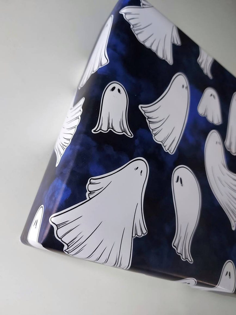 May include: A roll of wrapping paper with a dark blue background and a pattern of white cartoon ghosts. The ghosts have simple, rounded shapes and are scattered across the paper. The paper is partially unrolled, revealing the pattern.