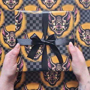May include: A black and yellow checkered wrapping paper with a repeating pattern of bats with pink ears and yellow eyes. The paper is tied with a black satin ribbon.