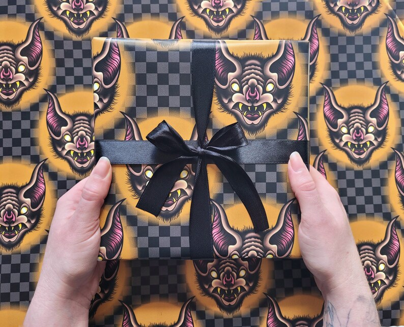 May include: A black and yellow checkered wrapping paper with a repeating pattern of bats with pink ears and yellow eyes. The paper is tied with a black satin ribbon.