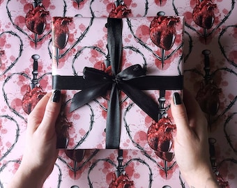 May include: Pink wrapping paper with a repeating pattern of black barbed wire, red hearts, and black daggers. The paper is wrapped around a gift box with a black satin ribbon.