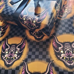 May include: Black and gold wrapping paper with a repeating pattern of cartoon bats with yellow eyes and pink ears. The bats have sharp teeth and are set against a black and white checkered background.