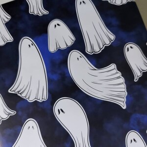 May include: White ghost illustrations on a dark blue background. The ghosts have simple, rounded shapes with two small eye details. The background has a watercolor-like effect, creating a spooky, Halloween-themed pattern.