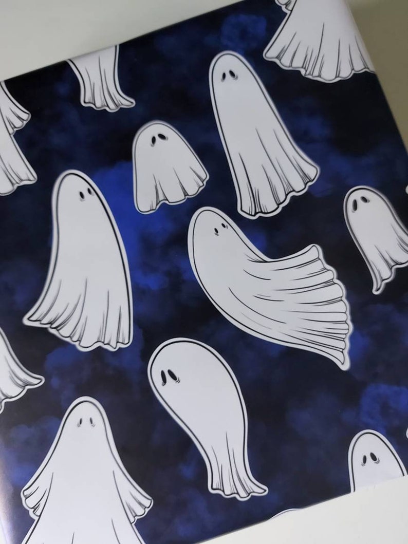 May include: White ghost illustrations on a dark blue background. The ghosts have simple, rounded shapes with two small eye details. The background has a watercolor-like effect, creating a spooky, Halloween-themed pattern.