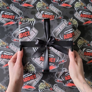 May include: Black and white wrapping paper with a horror movie theme. The paper features film reels, blood splatters, and the text "Horror Movie Addict". A black ribbon is tied around a roll of the wrapping paper.