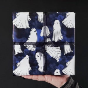 May include: A wrapped gift box featuring a repeating pattern of white cartoon ghosts on a dark blue background with a cloudy effect. The box is tied with a black ribbon, perfect for Halloween or spooky-themed gifts.