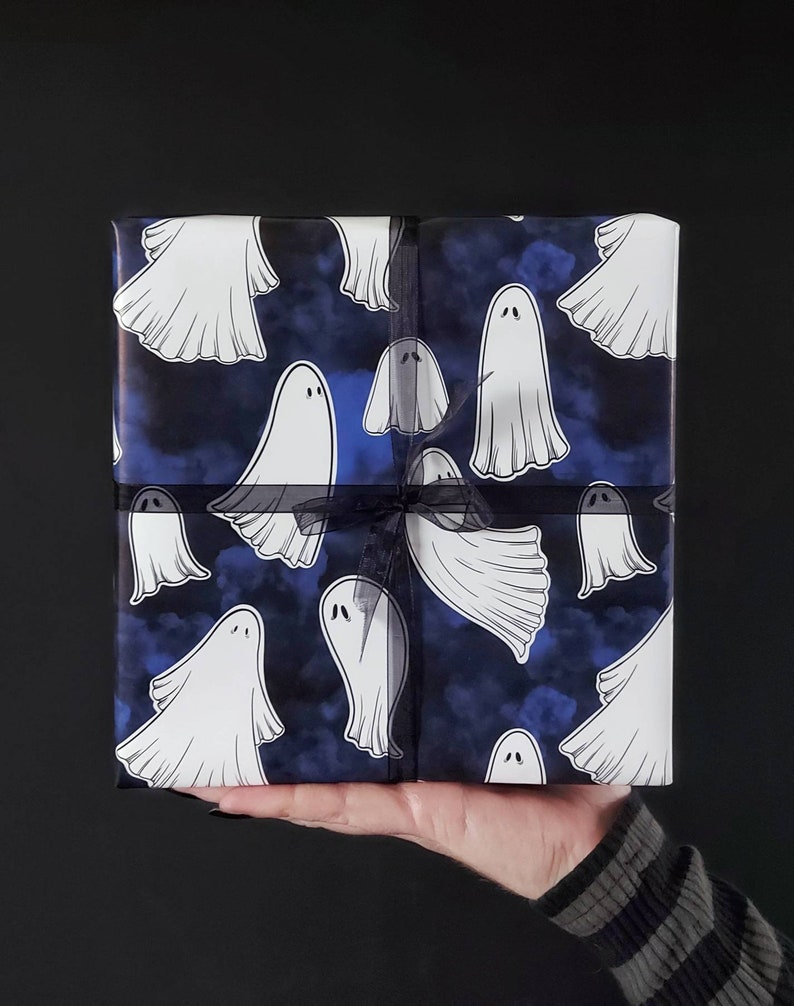 May include: A wrapped gift box featuring a repeating pattern of white cartoon ghosts on a dark blue background with a cloudy effect. The box is tied with a black ribbon, perfect for Halloween or spooky-themed gifts.