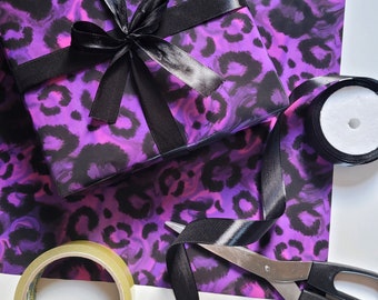 May include: Purple and black leopard print wrapping paper with a black satin ribbon tied in a bow. A roll of clear tape, a pair of scissors, and a roll of black ribbon are also visible.