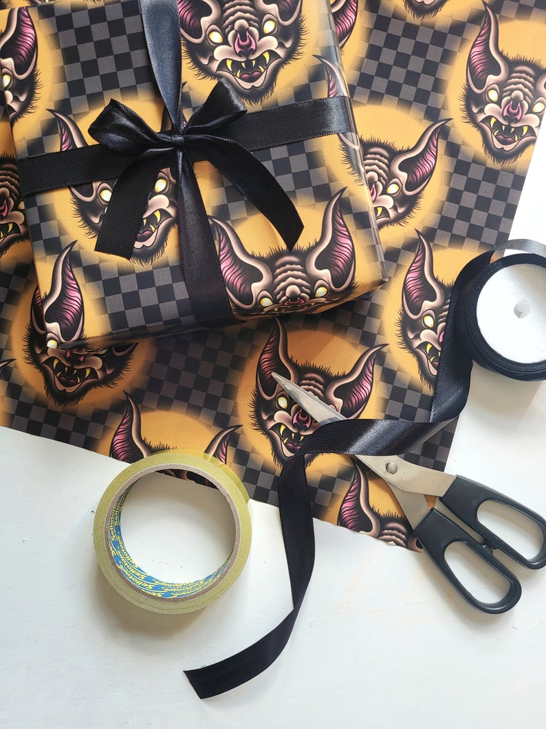 May include: A black and yellow checkered gift wrap with a repeating pattern of bats with pink accents. The gift wrap is wrapped around a box with a black ribbon and bow. There is also a roll of clear tape, a pair of scissors, and a roll of black ribbon.