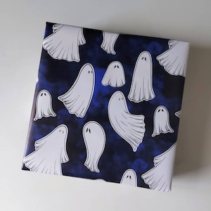 May include: Square gift wrap featuring a pattern of white cartoon ghosts on a dark blue background. The ghosts have simple, rounded shapes and are scattered across the surface. The background has a cloudy, textured appearance.