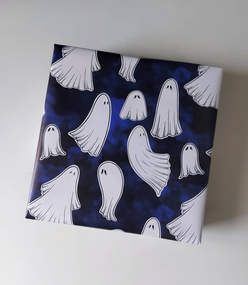 May include: Square gift wrap featuring a pattern of white cartoon ghosts on a dark blue background. The ghosts have simple, rounded shapes and are scattered across the surface. The background has a cloudy, textured appearance.