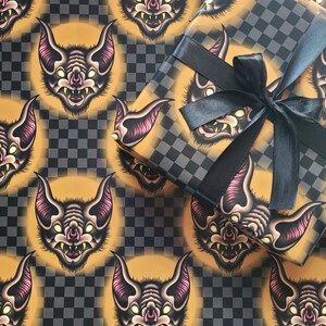 May include: A sheet of orange and black checkered wrapping paper with a repeating pattern of cartoon bats with pink ears and yellow eyes. The bats have sharp teeth and are facing forward.