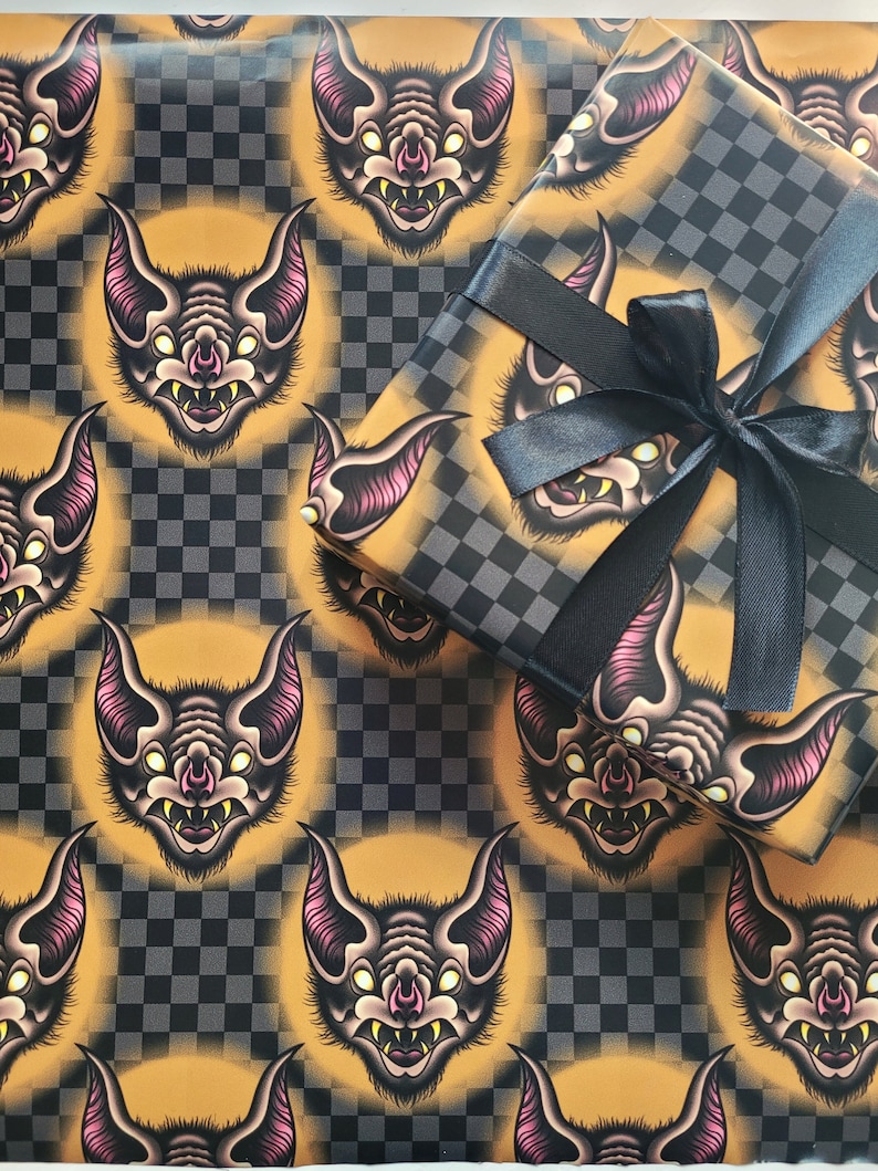 May include: A sheet of orange and black checkered wrapping paper with a repeating pattern of cartoon bats with pink ears and yellow eyes. The bats have sharp teeth and are facing forward.