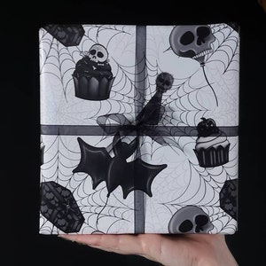 Peut inclure: Un papier cadeau blanc avec un motif de toile d'araign&eacute;e noir et blanc. Le papier pr&eacute;sente des illustrations noires et blanches de chauves-souris, de cercueils, de cupcakes et de cr&acirc;nes.