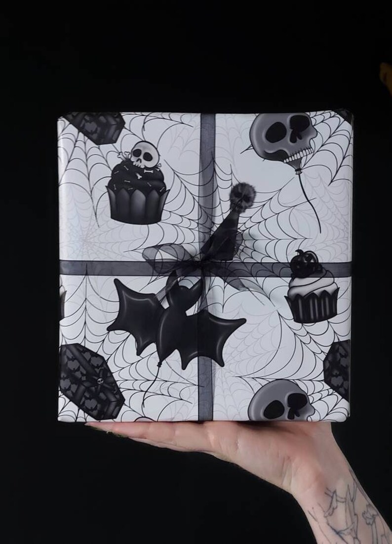 Peut inclure: Un papier cadeau blanc avec un motif de toile d'araign&eacute;e noir et blanc. Le papier pr&eacute;sente des illustrations noires et blanches de chauves-souris, de cercueils, de cupcakes et de cr&acirc;nes.