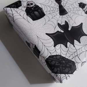 Peut inclure: Papier d'emballage noir et blanc avec un motif de toile d'araign&eacute;e et des illustrations sur le th&egrave;me d'Halloween, notamment des chauves-souris, des cr&acirc;nes, des cupcakes et des bo&icirc;tes cadeaux.