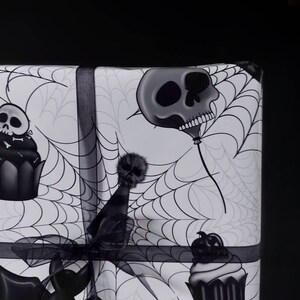 Peut inclure: Papier cadeau d'Halloween noir et blanc avec un motif de toile d'araign&eacute;e. Le papier pr&eacute;sente un ballon en forme de cr&acirc;ne, un cupcake surmont&eacute; d'un cr&acirc;ne et un cr&acirc;ne portant un chapeau de f&ecirc;te.