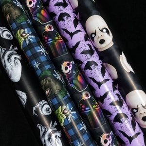 May include: Five rolls of wrapping paper with different spooky designs. The designs include a black and white anatomical heart, a blue and green plaid with a dinosaur, a purple background with bats, a black background with a rainbow cupcake, and a black background with a baby doll.