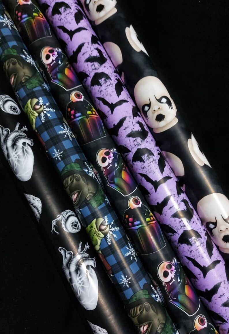 May include: Five rolls of wrapping paper with different spooky designs. The designs include a black and white anatomical heart, a blue and green plaid with a dinosaur, a purple background with bats, a black background with a rainbow cupcake, and a black background with a baby doll.