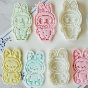 Puede incluir: Un conjunto de cortadores y sellos en relieve con forma de conejito en colores pastel, incluyendo verde, rosa, azul y amarillo. El texto "FOREST MONSTERS" se encuentra en la parte superior de la imagen, con "Cutter & embosser stamp set" debajo.