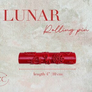 Puede incluir: Rodillo de pl&aacute;stico rojo con un dise&ntilde;o decorativo. El rodillo mide 10 cm de largo. El texto "LUNAR Rolling pin" est&aacute; por encima del rodillo. El texto "GCC" est&aacute; en un c&iacute;rculo debajo del rodillo.