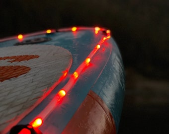 Könnte beinhalten: Ein blaues und orangefarbenes aufblasbares Paddleboard mit roten LED-Lichtern am Rand. Die Lichter sind eingeschaltet und leuchten hell.