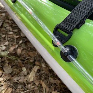 May include: A clear plastic tube with a red dot on it is attached to a black strap and a green kayak. The tube is secured to the kayak with a black plastic piece.
