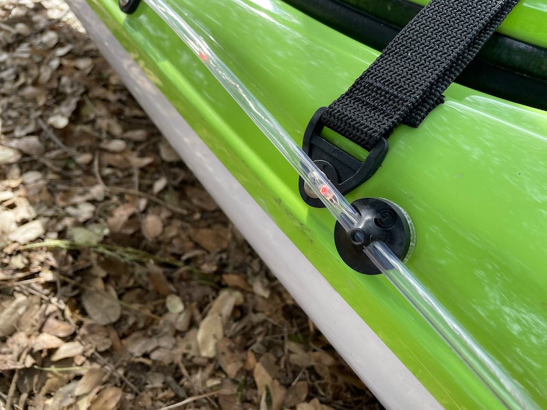 May include: A clear plastic tube with a red dot on it is attached to a black strap and a green kayak. The tube is secured to the kayak with a black plastic piece.