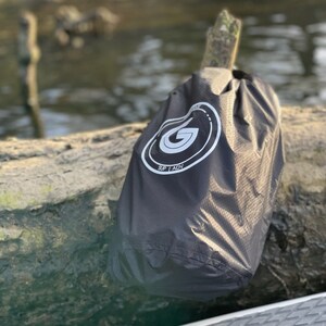 May include: Black waterproof dry bag with a white logo that says "SUP | ADV". The bag is sitting on a log near a body of water.