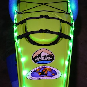 May include: A green kayak with a black strap and a sticker that says "Adventure Awaits" and "Church of the Double-Bladed Paddle". The kayak is decorated with green LED lights.