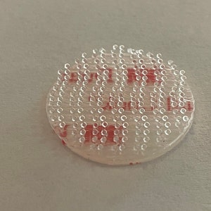 May include: A round, clear adhesive sticker with a red adhesive backing. The sticker has a grid of small, clear dots.
