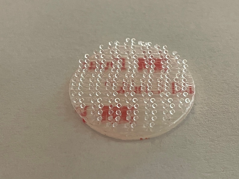 May include: A round, clear adhesive sticker with a red adhesive backing. The sticker has a grid of small, clear dots.