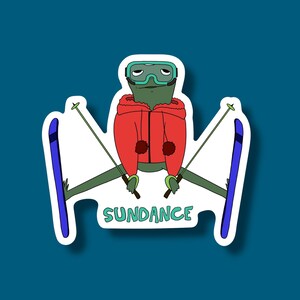 May include: A sticker featuring a cartoon frog wearing ski goggles and a red jacket, holding ski poles, and skis. The word "SUNDANCE" is written in green below the frog.