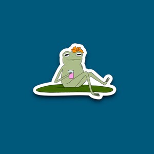 May include: A sticker featuring a cartoon frog relaxing on a lily pad. The frog is green with a flower on its head and is holding a pink drink. The background is a solid teal color.