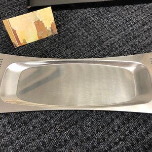 May include: A silver serving tray with a slightly curved shape and a raised edge. The tray has a smooth, polished surface and a decorative pattern on the sides.