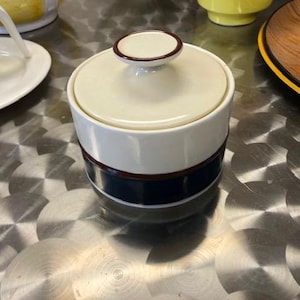 以下が含まれることがあります： 青と茶色の縁取りが施された白い陶器製の砂糖入れ。蓋の上には小さなつまみがある。