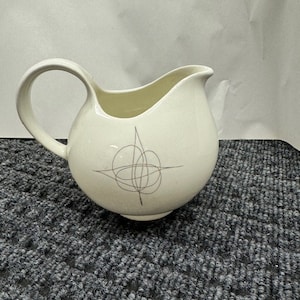 May include: A white ceramic creamer with a silver abstract design. The creamer has a rounded body and a curved handle.