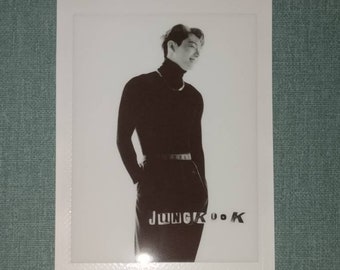 May include: Black and white photograph of a person wearing a black turtleneck sweater, black trousers, and a silver chain necklace. The photo is in a white frame with the text "JUNGKOOK" printed in black.