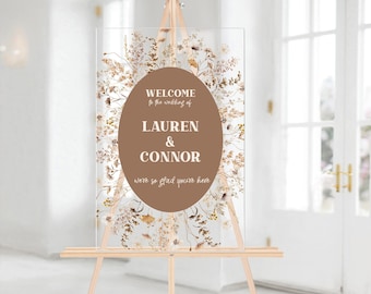 May include: Clear acrylic wedding welcome sign on a wooden easel. The sign features a brown oval with the names "Lauren & Connor" and the text "Welcome to the wedding of" and "we're so glad you're here". The sign is decorated with dried flowers.