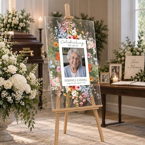 May include: A clear acrylic memorial sign on a wooden easel featuring a colorful floral border surrounding a portrait of an older person. The text reads Celebrating the life of Veronica Landoll, August 1, 1930 - April 9, 2022 in black font.