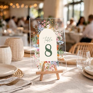 May include: Clear acrylic table number eight with floral design. The number eight is in a dark green color, and the word "Table" is written above it. The table number is displayed on a small wooden easel.