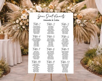 May include: A white wedding seating chart on a gold stand, with the text "Your Seat Awaits Amanda & Colin." The chart lists names under table numbers 1-12. The background features floral arrangements and outdoor decor.