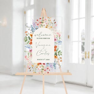 May include: A clear acrylic wedding welcome sign on a wooden easel. The sign features floral artwork and text that reads "welcome to our forever" and the names "Vivienne & Carlos" with the date "August 5th, 2028".
