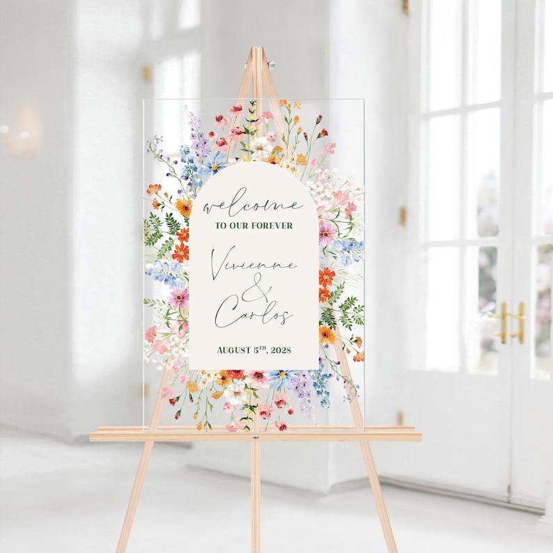 May include: A clear acrylic wedding welcome sign on a wooden easel. The sign features floral artwork and text that reads "welcome to our forever" and the names "Vivienne & Carlos" with the date "August 5th, 2028".