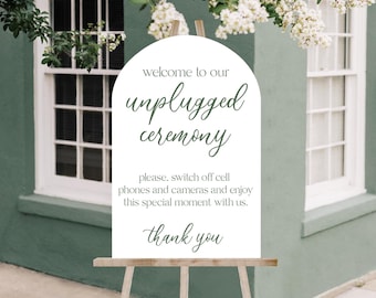 Op de afbeelding: Een wit bord op een houten ezel met de tekst "welcome to our unplugged ceremony" in groen schrift. Het bord vraagt gasten om telefoons en camera's uit te schakelen. De achtergrond is een groene muur en witte bloemen.